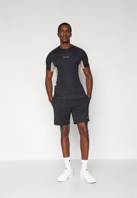 Μαύρο αθλητικό t-shirt με μπεζ πλαϊνές επιφάνειες, κοντά μανίκια και μικρό λογότυπο στο στήθος. Συνδυάζεται με μαύρο σορτς και λευκά αθλητικά παπούτσια.