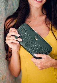 Green leather wallet featuring a crocodile pattern, zip closure, and a gold emblem. Held in a hand with neutral nail polish.