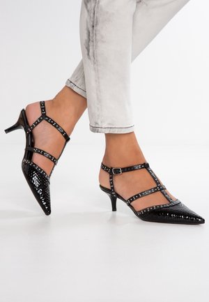 Zwarte schoenen met lage hak en puntige neus met gespikkelde bandjes, gedragen met lichtgrijze opgerolde jeans op een witte vloer.