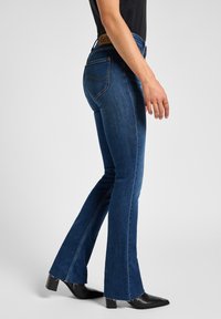 Jeans acampanados de denim azul con una cintura de altura media, costuras visibles en los bolsillos traseros y dobladillo deshilachado. Combinados con botas negras de punta afilada.