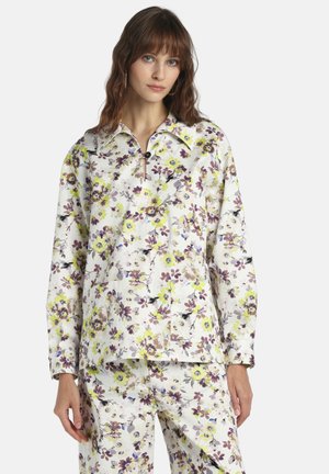 Kobieta ubrana w białą koszulę z długim rękawem i spodnie w kwiatowy wzór w kolorach fioletowym, żółtym i brązowym, stojąca na jednolitym tle.