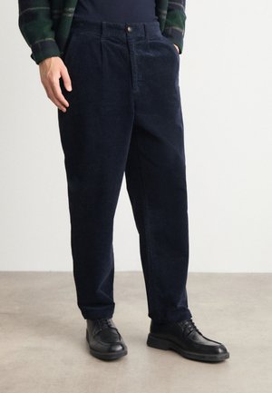 Marineblauwe corduroy broek met een ontspannen pasvorm, met een knoopsluiting en voorplooien. Gecombineerd met zwarte veterschoenen op een lichtgrijze vloer.