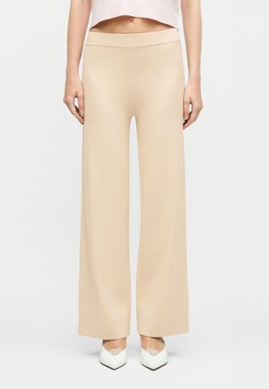 Nő bézs színű, bőszárú nadrágban és fehér, hegyes orrú cipőben, világos semleges háttér előtt áll.