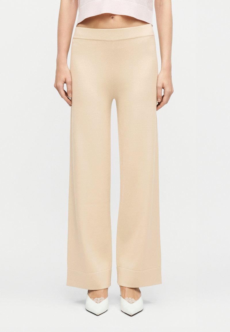 Γυναίκα που φοράει μπεζ παντελόνι καμπάνα και λευκά μυτερά παπούτσια, στέκεται μπροστά σε απλό φωτεινό φόντο.