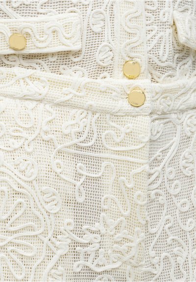 Tissu en maille blanche avec une broderie en relief complexe et trois boutons ronds en or disposés verticalement sur le vêtement.