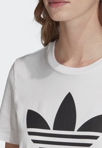 Witte katoenen T-shirt met een zwart trefoil-logo met drie bladeren en horizontale strepen op de borst, korte mouwen en een ronde hals.
