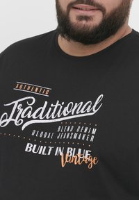Crna pamučna majica s bijelim i narančastim natpisima "Autentično Tradicionalno" i "Izrađeno u Plavoj Vintage," s okruglim izrezom.