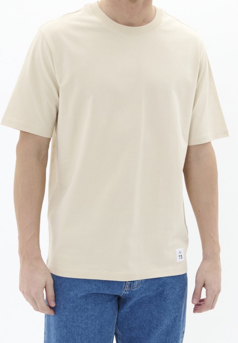 T-shirt beige à manches courtes en tissu lisse, avec un col rond et une étiquette sur le côté inférieur indiquant la taille et la marque.