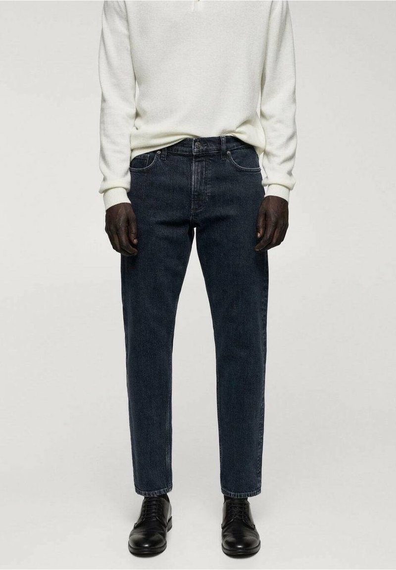 Personne portant un jean droit bleu foncé, un pull blanc à manches longues et des chaussures noires à lacets, debout contre un fond uni.