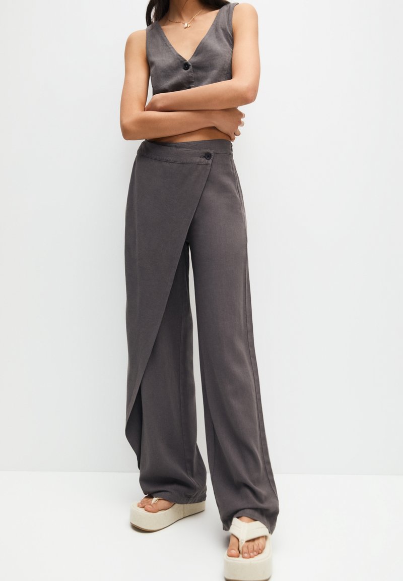 Top corto sin mangas en gris con diseño de envoltura y pantalones de pierna ancha a juego con detalle de botones, combinados con sandalias de plataforma blancas sobre un fondo claro.