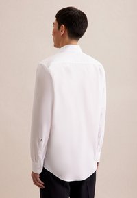 Chemise blanche à manches longues avec un col pointu, texture lisse, ourlet arrondi et poignets à boutons, portée par une personne vue de dos.