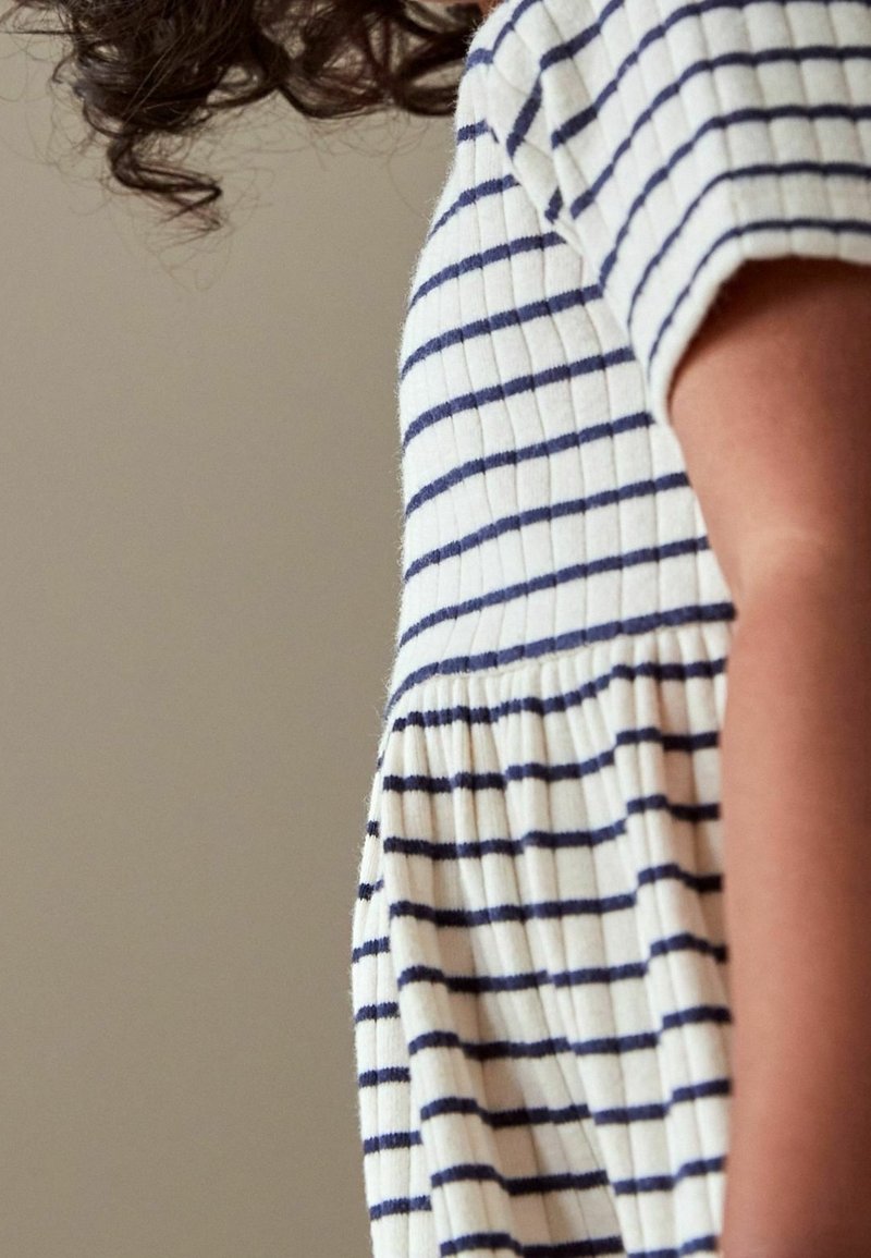 Pasiasta sukienka w białym i granatowym kolorze, z prążkowaną fakturą, krótkimi rękawami i marszczoną spódnicą. Skupienie na widoku z boku sukienki.