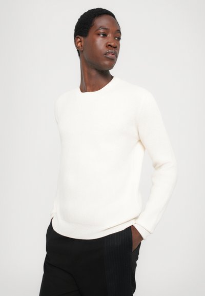 White ribbed sweater with a crew neckline, long sleeves, and a relaxed fit, paired with black pants featuring pinstripes.