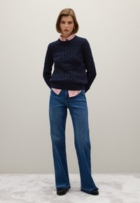 Marineblaue Zopfmuster-Strickjacke über einem rosa-weiß gestreiften Hemd, kombiniert mit blauen Schlagjeans. Die Socken sind schwarz und haben eine strukturierte Oberfläche.
