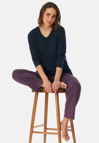 Top a maniche lunghe V in blu navy abbinato a pantaloni da pigiama a righe viola, con una vestibilità comoda e una texture morbida, modella seduta su uno sgabello.