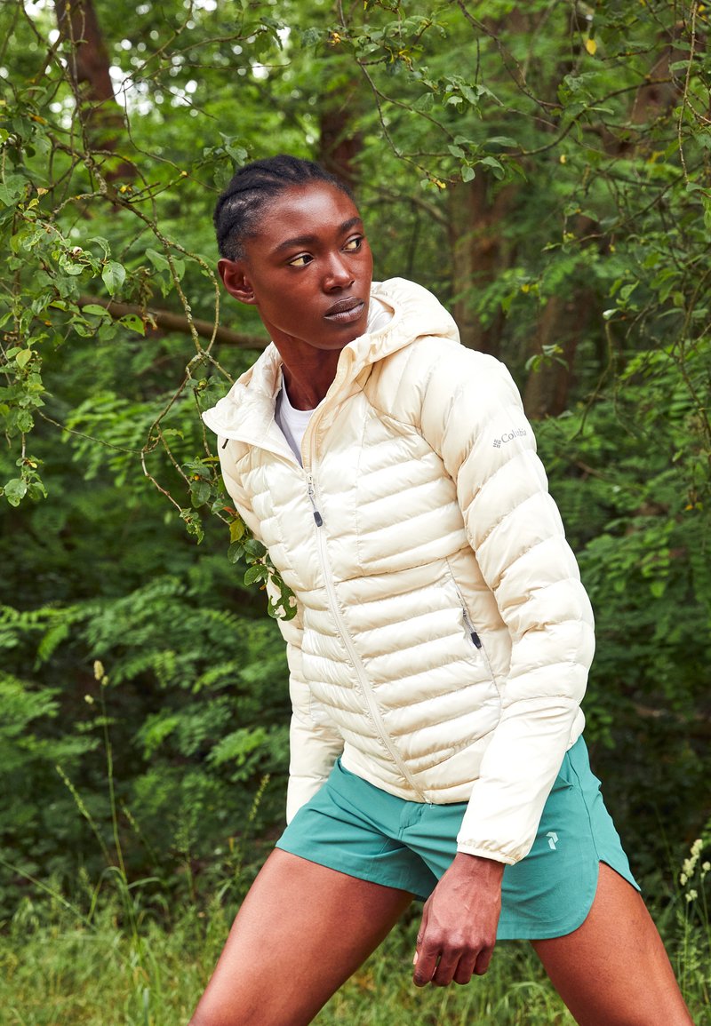 Chaqueta ligera y acolchada de color blanco roto con capucha, cuenta con costuras horizontales, cierre delantero con cremallera y bolsillos laterales, sobre un fondo verde con hojas.