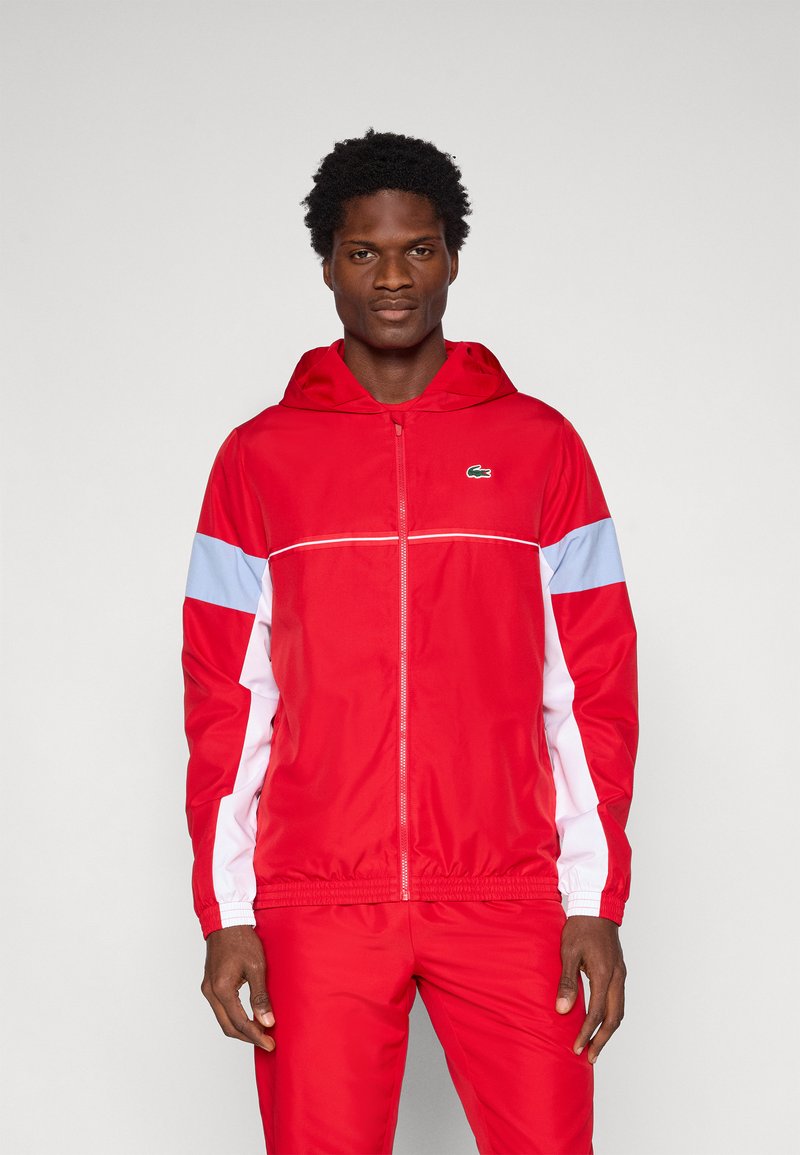 Κόκκινο ελαφρύ μπουφάν με καπέλο, με λευκές και ανοιχτό μπλε πινελιές, διαθέτοντας μπροστινό φερμουάρ και ελαστικές μανσέτες. Λογότυπο Lacoste στο στήθος.