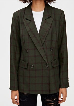 Donkergroene dubbelgeknoopte blazer met bordeauxrood ruitjespatroon, gedragen over een wit topje en gecombineerd met zwarte geripte jeans.