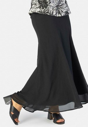 Persona con una falda larga negra y sandalias negras de tacón cuadrado con tiras cruzadas, de pie contra un fondo liso.