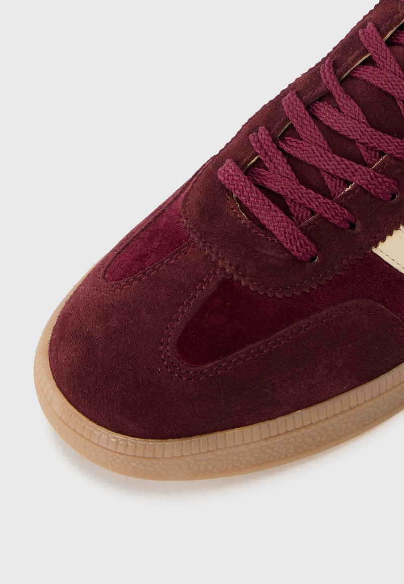 Burgunderfarbene Wildleder-Sneaker mit strukturiertem Obermaterial, flachen Schnürsenkeln, auffälliger Seitenpartie und einer Gummigummisohle, die sich auf sportliche Designelemente konzentriert.