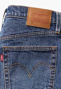 Džinsai su mėlynai tonu, turintys rudą odinį ženkliuką, vario siuvinėjimą ir raudoną Levi's etiketę ant užpakalinio kišenės su V formos siūle.