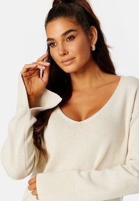 Pullover a maglia beige con scollatura a V profonda, polsini a coste e vestibilità rilassata. Presenta maniche lunghe e una texture morbida.