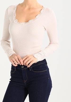 Vrouw draagt een lichtroze geribd shirt met lange mouwen en kanten rand aan de halslijn en manchetten, gecombineerd met donkerblauwe high waisted jeans.