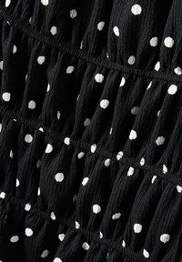 Czarna teksturowana tkanina w białe kropeczki, z falbaniastymi sekcjami, tworzącymi marszczenia. Podkreśla grę wzorów i kontrastów.