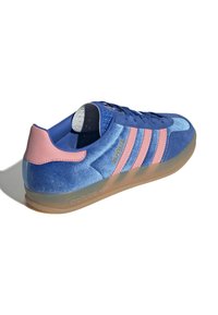 Μπλε σουέτ αθλητικά παπούτσια με ροζ λεπτομέρειες, διαθέτοντας τρεις ροζ ρίγες, υφασμένη φτέρνα και διαφανή σόλα από καουτσούκ. Χρυσό λογότυπο "Gazelle" εμφανίζεται.