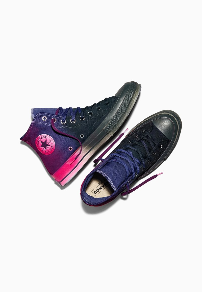 Hohe schwarze und lila Converse All Star Sneaker mit pinken Akzenten, ein Schuh flach und einer schräg, Schnürsenkel teilweise geöffnet auf weißem Hintergrund.