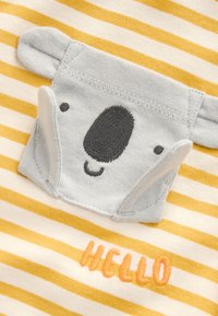 Aplicación de cara de koala con orejas plegables sobre tela a rayas amarilla y blanca con texto bordado en naranja que dice hola.