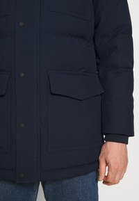 Hombre con chaqueta acolchada azul marino con bolsillos de solapa y botones de presión, combinada con vaqueros azules, mano visible en el lado.