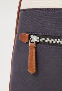 Détail d'un sac en tissu marine avec une fermeture éclair argentée, un tirant en cuir brun, et une surface lisse avec des accents beige clair.