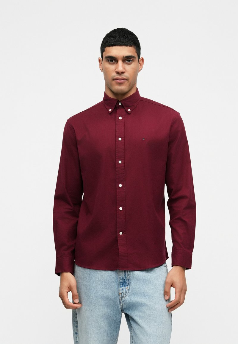 Bordó gombos ing gallérral és hosszú ujjakkal, fehér gombokkal és egy kis logóval a mellkason. Világoskék farmernadrággal viselve.