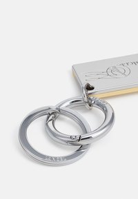 Anneaux en métal argenté attachés à une étiquette de porte-clés rectangulaire avec des bords dorés, gravée du nom de la marque "Lancel".
