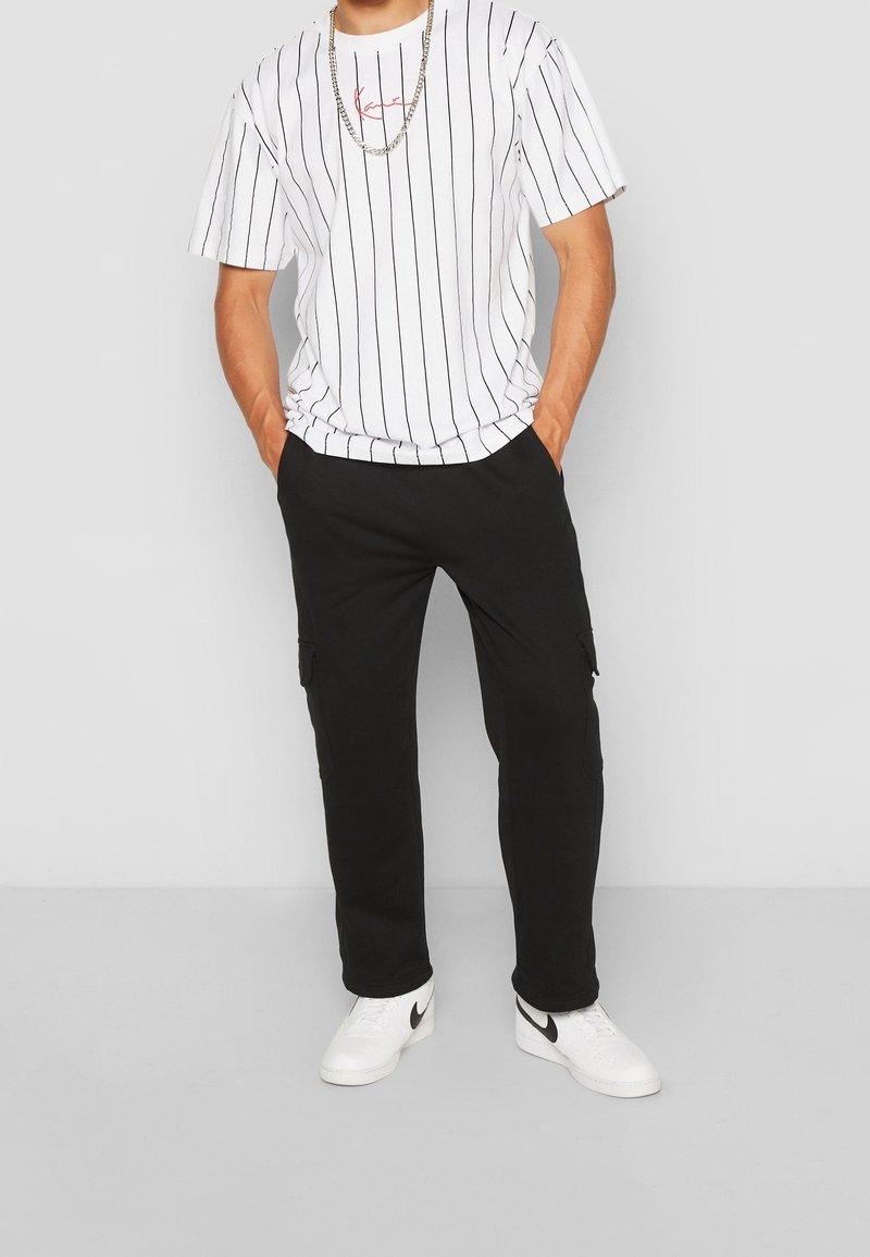 Mężczyzna w białej koszulce w paski, czarnych spodniach cargo, białych sneakerach Nike i srebrnym łańcuszku, stojący z rękami w kieszeniach.