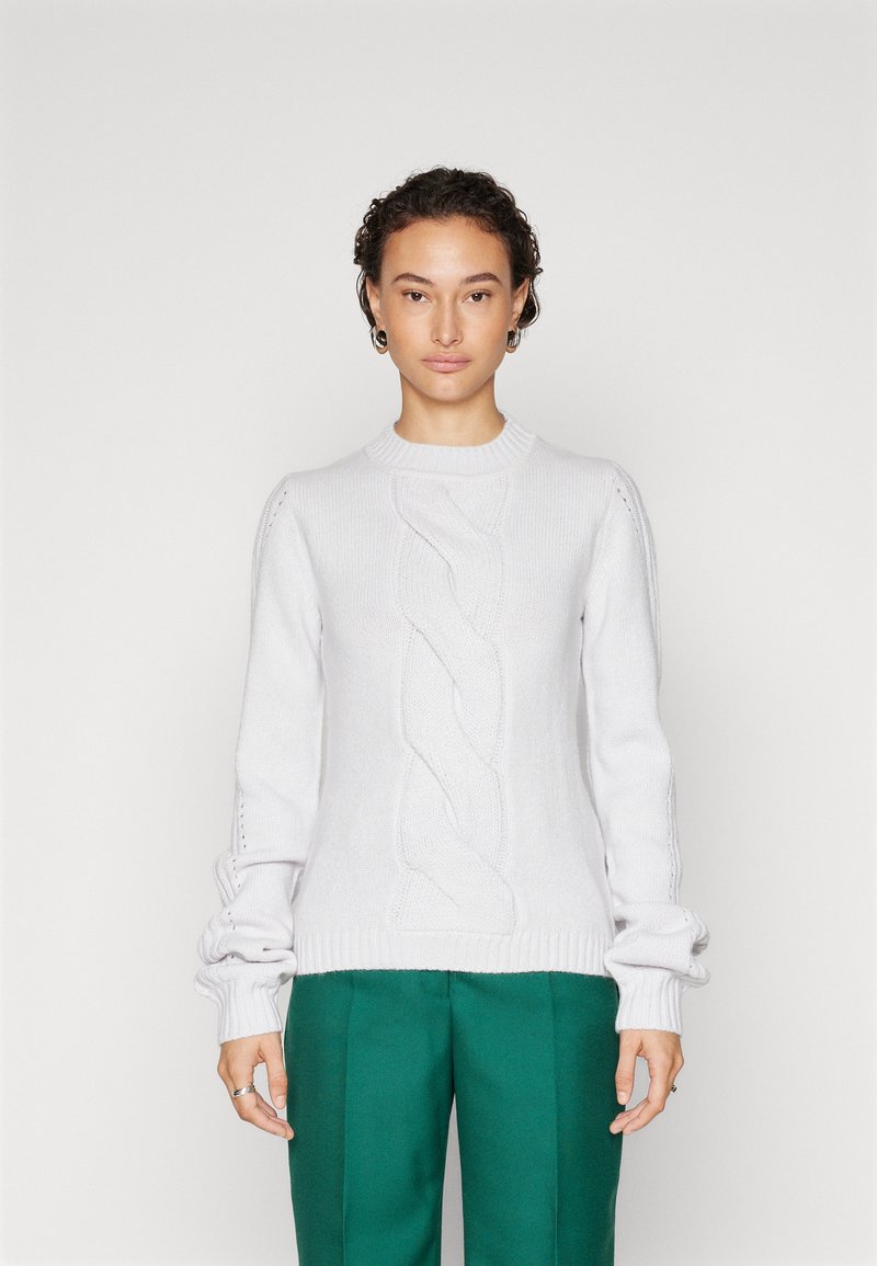 Donna con capelli ricci corti, che indossa un maglione bianco a treccia e pantaloni verdi, in piedi contro uno sfondo chiaro e semplice.