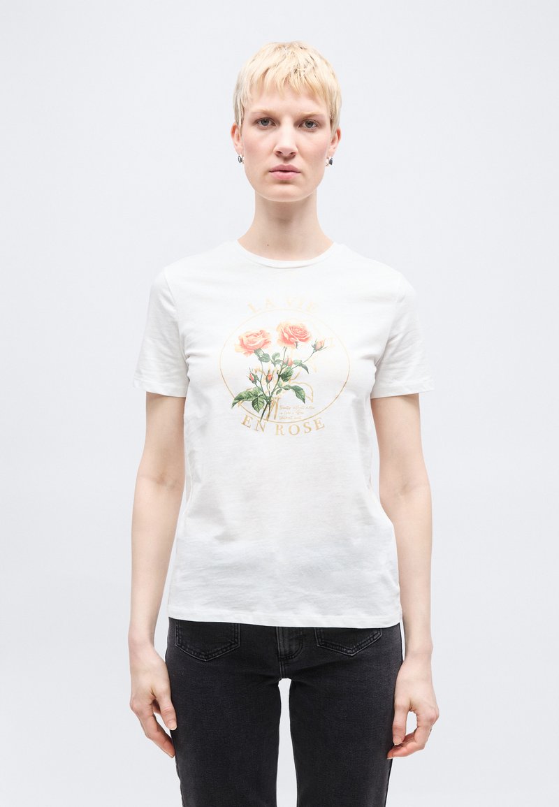 Άτομο με κοντά ξανθά μαλλιά που φοράει λευκό μπλουζάκι με γραφικό τριαντάφυλλο και κείμενο «La Vie En Rose», συνδυασμένο με μαύρα τζιν.