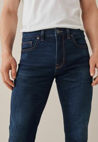 Donkerblauwe spijkerbroek met een getailleerd ontwerp, voorzien van oranje stiksels, vijf zakken en een knoopsluiting bij de taille.