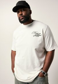 Man met baard, gekleed in een zwarte pet, wit shirt met Franse tekst op de borst, handen in de zakken, polshorloge en kettinghalsketting tegen een effen achtergrond.