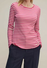 Vrouw in een lichtroze en witte gestreepte langemouwen shirt met een gekruld zoom, met haar handen in de zakken van donkerblauwe broek, tegen een effen achtergrond.