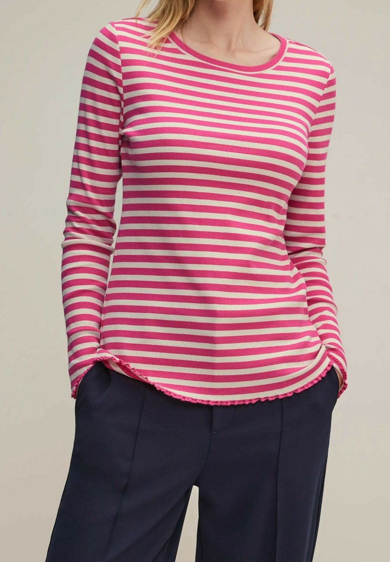 Vrouw in een lichtroze en witte gestreepte langemouwen shirt met een gekruld zoom, met haar handen in de zakken van donkerblauwe broek, tegen een effen achtergrond.