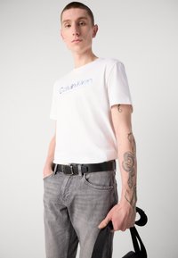 Balta kokvilnas T krekliņa ar zilo "Calvin Klein" logotipu, apvienojot ar pelēkiem džinsu biksēm un melnu ādas jostu. Modelis tur rūtiņu.