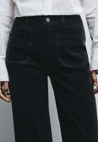 Donkerblauwe corduroy broek met een rechte pijp, voorzien van twee voorzakken en een zilveren knoop in de taille.