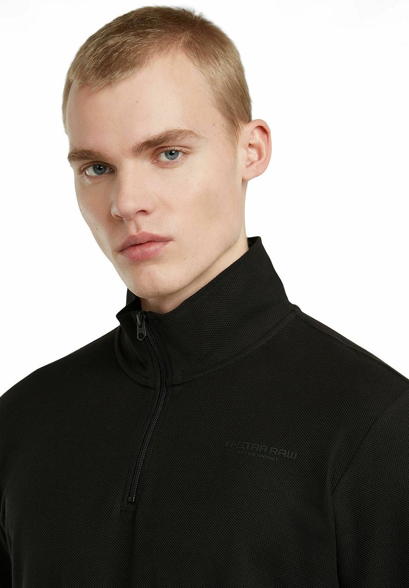 Schwarze, mit Reißverschluss versehene Jacke aus strukturiertem Stoff, mit hohem Kragen und dezentem Logo-Akzent. Die eng anliegende Passform erhöht die Wärme.