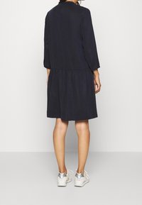 Una donna indossa un vestito nero al ginocchio con maniche a tre quarti e sneakers bianche, in piedi davanti a uno sfondo bianco uniforme.