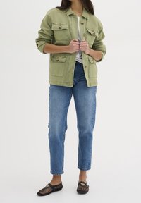 Woman wearing light green buttoned jacket over white shirt, blue jeans, and black mesh flat shoes, standing against a plain background.