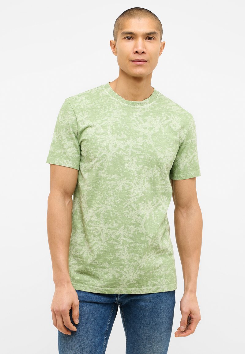 Grünes kurzärmeliges T-Shirt mit einem subtilen Blattmuster, rundem Ausschnitt und weicher Textur. Kombiniert mit blauen Denim-Jeans.