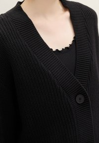 Musta neulottu neuletakki, jossa syvä V-pääntie, ribbineulos ja yksittäinen nappikiinnitys. Päällä musta röyhelöinen hihaton toppi.