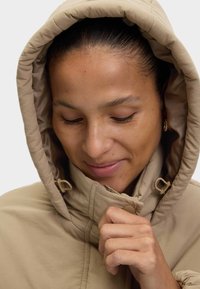 Beige hættetrøje med høj krave, der har en glat tekstur og knap- og løslukninger, med fokus på den øverste del og hænderne.